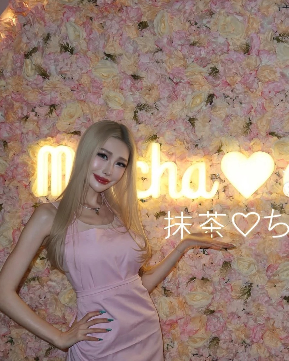 Woman in a pink dress posing in front of the glowing Matcha-chan neon sign and pink flower wall